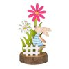 Figurina decor pentru masa de Paste, iepuras pe stativ din lemn, gard alb, iarba verde si flori multicolor de primavara, inaltime 13.5 cm